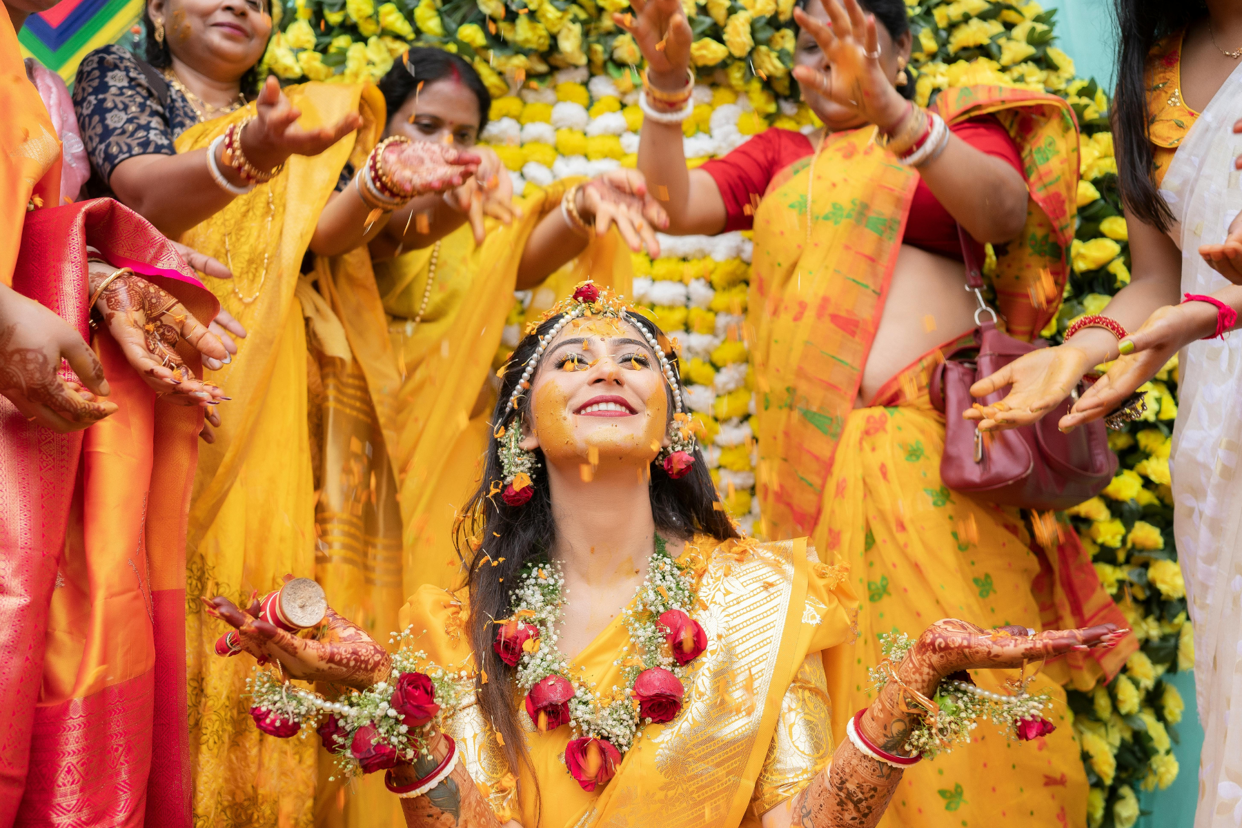 Bride enjoying her Haldi ceremony surrounded by women in yellow sarees, smiling with flower petals showered during Indian wedding ritual in Delhi NCR.