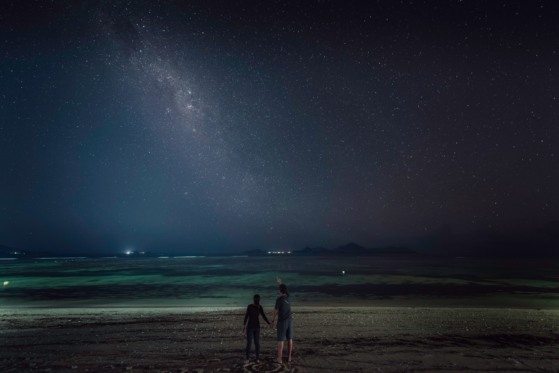 Two people stand hand in hand on a quiet beach at night, facing the ocean. One person points toward the sky, which is filled with countless stars and the glowing arc of the Milky Way. The scene is calm and serene, with faint lights visible on the distant horizon and silhouettes of islands in the background.